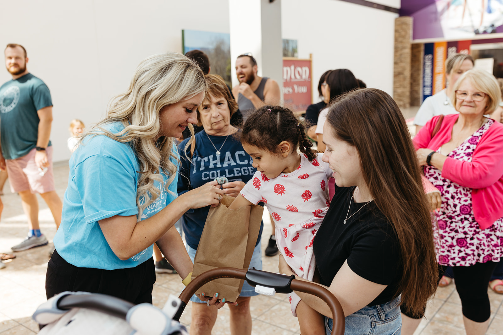 A smiling woman hands a small gift bag to a young child being held by her mother during a community fundraiser event, surrounded by families and supporters